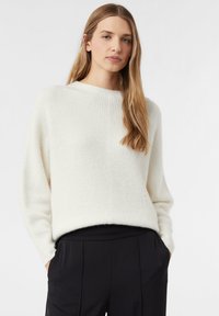 Cremefarbener, gerippter Strickpullover mit Rundhalsausschnitt und überschnittenen Schultern, kombiniert mit hoch taillierten schwarzen Plisseehosen. Weiche Textur.