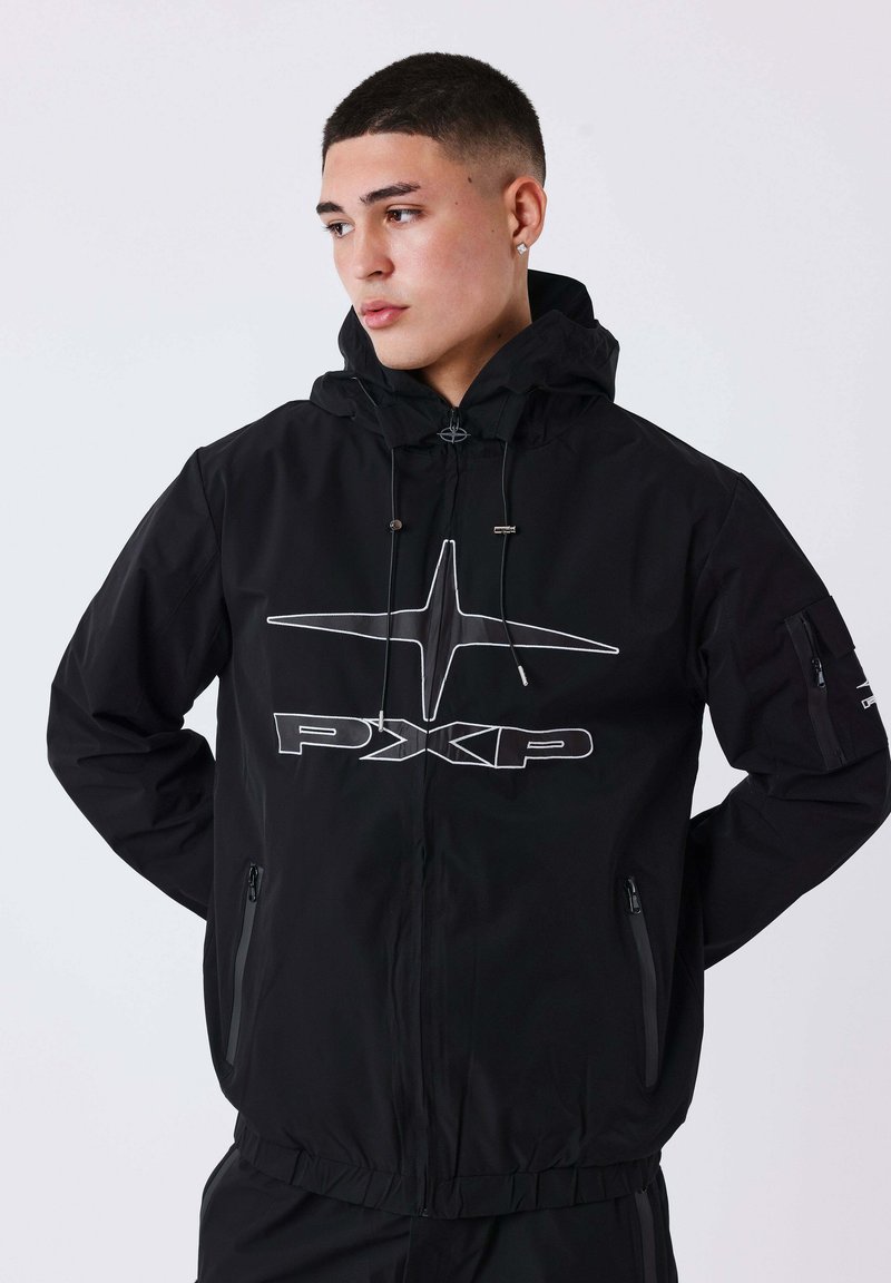 Giovane uomo con i capelli corti che indossa una giacca nera con cappuccio e un grande logo bianco "PXP" e zip frontale, che guarda di lato.