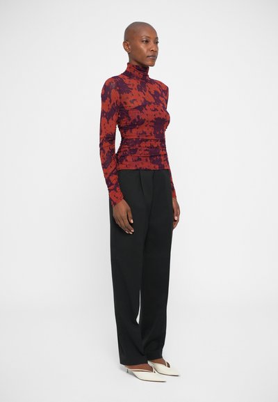 Haut à manches longues rouge et noir à motifs avec un col montant, associé à un pantalon noir ajusté et des chaussures blanches à bout pointu. Texture lisse.