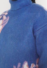 Niebieski sweter z dzianiny z wysokim golfem, posiadający kwiatowy wzór w delikatnym różu i bieli na dole oraz teksturowane wykończenie.