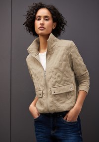Beige zip-jacka med strukturerad design, hög krage och två framfickor. Buren över en vit skjorta, i kombination med blå denimjeans.