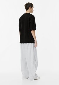 Mand står i siden iført en løs sort T-shirt, brede lysegrå bukser og hvide sneakers foran en ensfarvet hvid baggrund.