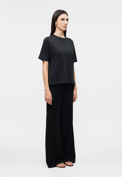 Γυναίκα που στέκεται, φορώντας ένα άνετο μαύρο μπλουζάκι με κοντά μανίκια, μαύρο παντελόνι με φαρδιά μπατζάκια και καφέ επίπεδα σανδάλια, με φόντο λευκό και απλό.