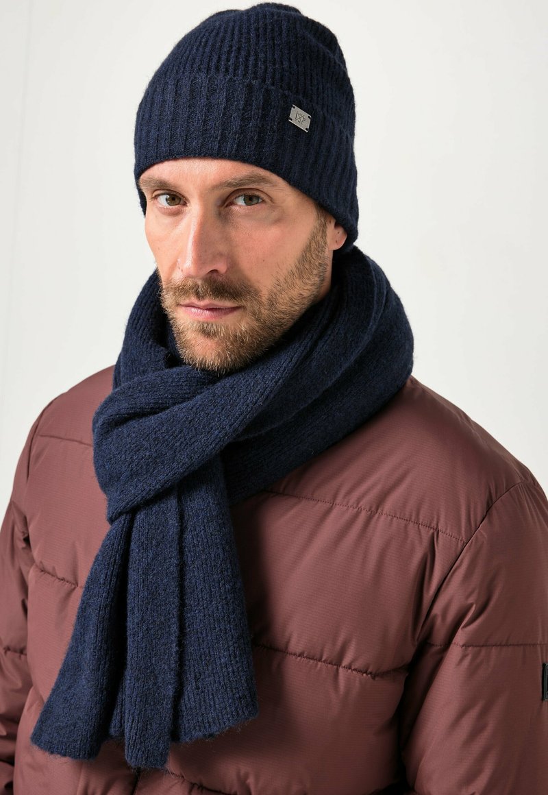 Navy ribbed knit beanie and matching scarf, both made from textured fabric, worn over a brown puffy coat with a visible logo accent.