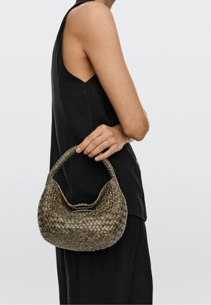 Persoon in zwart mouwloos topje houdt geweven, gebogen handtas met korte handgreep tegen een effen lichte achtergrond.