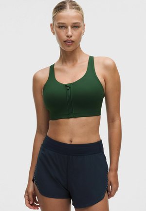 Femme portant un soutien-gorge de sport vert foncé à fermeture éclair sur le devant et un short de sport bleu marine, debout devant un fond clair uni.
