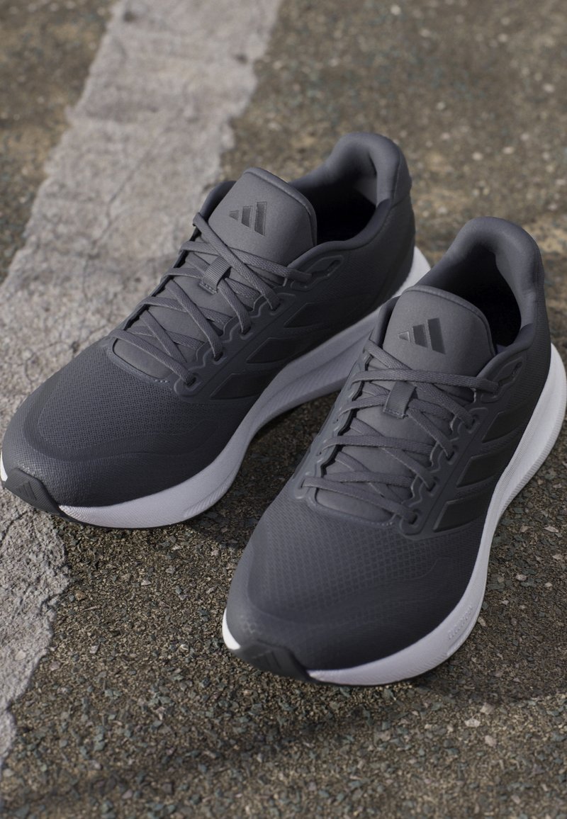 Paire de chaussures de sport gris foncé avec semelles blanches, lacets et logo discret sur les languettes, posées sur une surface asphaltée.