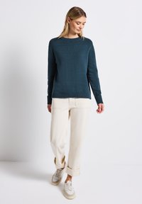 Blauer gestrickter Pullover mit rundem Halsausschnitt, gerippten Bündchen und Saum; kombiniert mit cremefarbener, lässiger Hose und neutralen Sneakern.