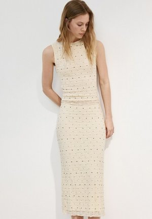 Kvinde iført en ærmeløs, cremefarvet, tekstureret midikjole med små sorte prikker, stående mod en hvid væg og kigger ned.