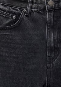 Gros plan sur un jean en denim noir montrant la poche avant, la passants de ceinture, la couture et un bouton en métal avec gravure de la marque.