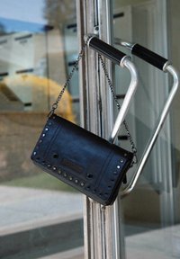Zwarte leren handtas met zilveren studs en kettinghengsel, met een gestructureerd paneel en een ritssluiting, hangend aan een deurhendel.