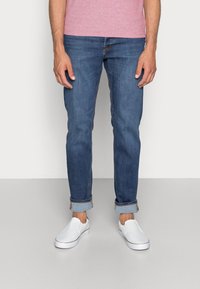 Camiseta rosa claro para hombre, jeans de denim azul con dobladillos enrollados y zapatillas blancas sin cordones, con una textura suave y un diseño clásico.