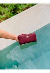 Maroon leather wallet with a textured surface, featuring a gold logo and a zip closure, held above a pool's edge. Water droplets visible.