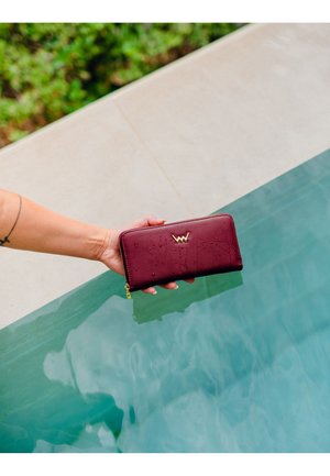 Maroon leather wallet with a textured surface, featuring a gold logo and a zip closure, held above a pool's edge. Water droplets visible.