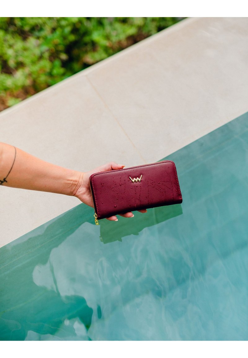 Maroon leather wallet with a textured surface, featuring a gold logo and a zip closure, held above a pool's edge. Water droplets visible.