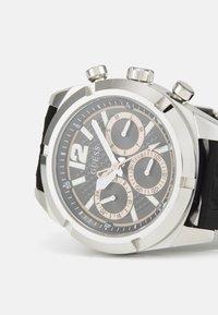 Reloj de acero inoxidable plateado con correa de goma negra. Presenta una esfera negra texturizada con tres subesferas y números en negrita.