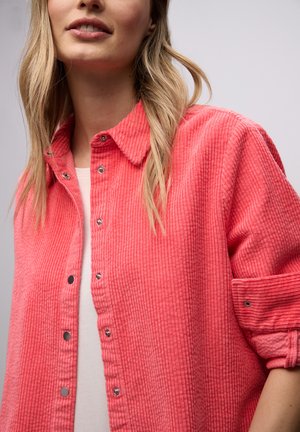 Femme portant une chemise en velours côtelé rose corail à boutons, par-dessus un haut blanc, avec les manches retroussées et les cheveux blonds lâchés.