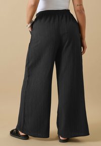 Czarne spodnie szerokie z teksturowanej tkaniny, z elastycznym paskiem i kieszeniami po bokach, w połączeniu z czarnymi klapkami.