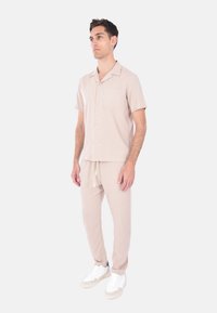 Camicia beige a maniche corte con tasca frontale, abbinata a pantaloni coordinati. Entrambi sono realizzati in un tessuto leggero. Le sneakers bianche completano il look.
