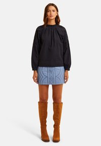 Blusa nera a maniche lunghe con scollatura arricciata, abbinata a una gonna azzurra chiara con motivi in rilievo, e stivali al ginocchio in suede color cammello.