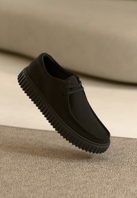 Sorte hverdagssko med tykk, ribbet såle som svever over en beige teksturert bakgrunn, vinklet for å vise side- og toppdetaljer.
