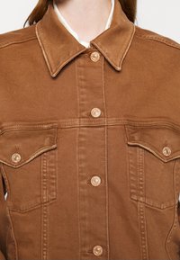 Veste en denim marron avec un col, deux poches avant et des coutures visibles. Dotée de boutons pression dorés et d'une texture lisse.