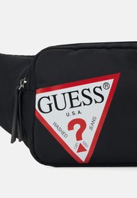 Černý nylonový bum bag s červeným a bílým trojúhelníkovým logem Guess, které obsahuje text a otazník. Obsahuje kapsu na zip.
