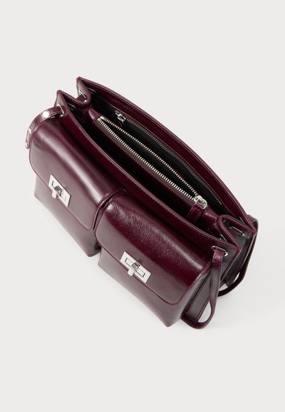 Σκούρα μοβ δερμάτινη τσάντα χειρός με δύο εμπρόσθιες τσέπες, ασημένιες κλειδαριές με περιστροφή και επάνω φερμουάρ, φαίνεται από πλάγια επάνω όψη.