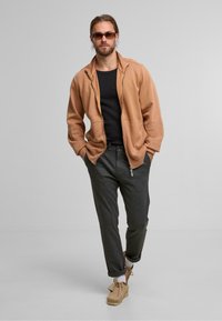 Beige Zip-Hoodie über einem schwarzen Oberteil, kombiniert mit dunkelgrauen, schmal geschnittenen Hosen und beigen Slipschuhen. Die Brille verleiht ein markantes Element.