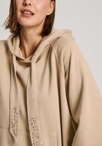 Sudadera con capucha beige de textura suave, con un bolsillo frontal y tachuelas decorativas en los cordones. Diseño casual y oversize.