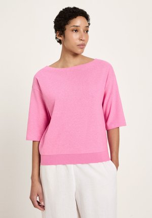 Woman with short curly hair wearing a loose pink short-sleeve top and white pants, looking to the side against a plain background.