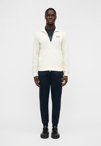 Ανδρικό μπουφάν με φερμουάρ σε κρεμ απόχρωση, μαλακό υλικό· συνδυασμένο με μπλε σκούρες αθλητικές φόρμες με λογότυπο EA7 και μαύρα αθλητικά παπούτσια.