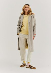 Svetlošedý vlněný kabát s béžovými knoflíky, žltý mikina s vyrazeným textom, béžové nohavice a tenisky s leopardím vzorom a viacfarebnými ponožkami.