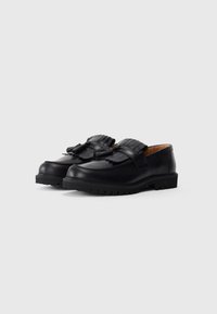 Une paire de mocassins en cuir noir avec des semelles épaisses, des détails en franges et à pompons, positionnée sur un fond gris clair uni.