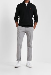 Svart zip-tröja, ljusgrå chinos och vita sneakers. Tröjan har en logotyp på bröstet; chinosen har en ren skärning.