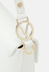 Weiße Lederhandtasche mit einem goldenen, runden Hardware-Detail und einem "V"-Akzent sowie einem flachen Tragegurt mit Druckknopfverschluss.