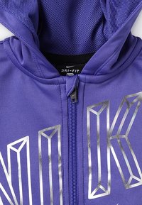 Lilla Nike Dri-FIT hættetrøje med fuld lynlås og reflekterende sølvgeometriske bogstaver på forsiden, med en mesh-foring i hætten.