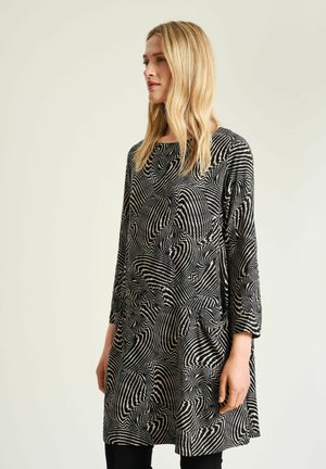 Femme aux longs cheveux blonds portant une robe noire et blanche à manches longues avec des motifs abstraits tourbillonnants sur un fond clair.