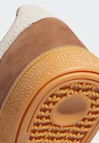 Hnedý zamšový zvršok so štruktúrovaným krémovým detailom, s dekoratívnou oranžovou gumovou podrážkou so vzorom a kruhovou centrálnou vložkou.