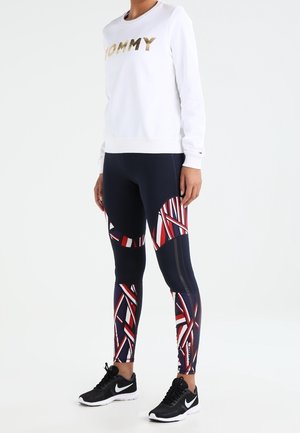 Biała bluza z napisem "TOMMY" w złotym kolorze, zestawiona z granatowymi legginsami z geometrycznym wzorem w czerwono-biało-niebieskie kolory oraz czarnymi butami sportowymi.