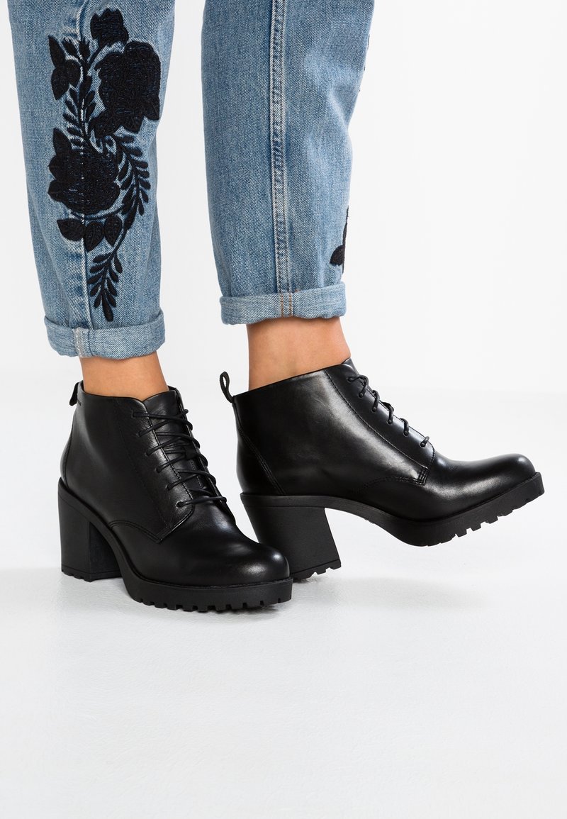 Botas de cuero negro hasta el tobillo con un tacón grueso, diseño de cordones y suela texturizada. Llevadas con jeans azul arremangados que presentan bordados florales en negro.