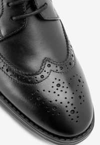 Chaussure habillée en cuir noir avec une finition lisse, un design à bouts arrondis, des détails de brogue et des perforations circulaires au niveau des orteils.