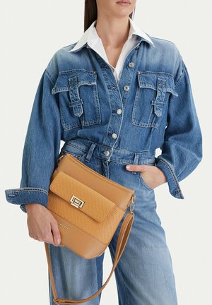 Mujer con chaqueta de mezclilla azul y vaqueros sostiene un bolso bandolera marrón claro con solapa tejida y cierre metálico.