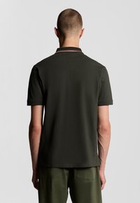 Mörkgrön polotröja med en kontrasterande randig krage. Korta ärmar och en slät textur. Synliga sömmar vid nederkanten.