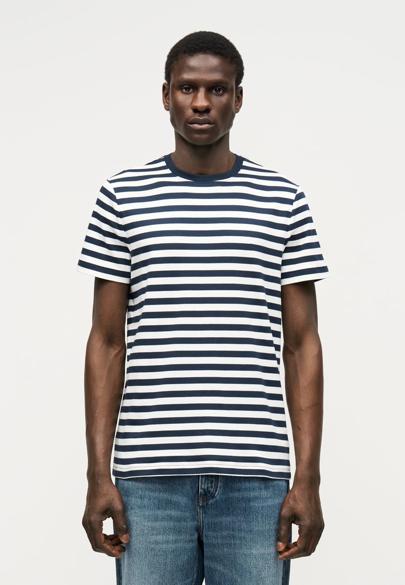 Άνδρας που στέκεται και κοιτά προς τα εμπρός φορώντας μπλε-σκούρο και λευκό οριζόντια ριγωτό t-shirt και μπλε τζιν μπροστά από απλό φόντο.