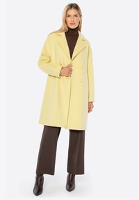 Abrigo amarillo de doble botonadura, hecho de un tejido suave, con cuello muesca y grandes botones. Combinado con un suéter de cuello alto marrón y pantalones.