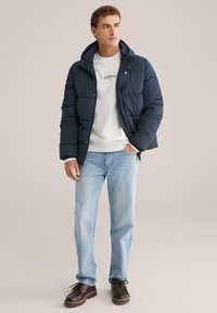 Mörkblå puffjacka med huva, knappstängning, bärs över en ljusgrå sweatshirt och ljusblå jeans, kompletterad med mörka snörskor.