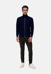 Navy ribbed zip-up sweater with high collar, paired with dark pants and white sneakers. The design features a fitted silhouette and smooth fabric.