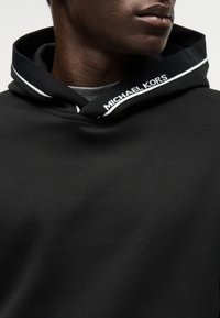 Lähivõte inimesest, kes kannab musta kapuutsiga pusa, millel on kapuutsi äärel kiri „Michael Kors“ halli särkkaeluse kõrval.