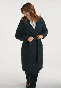 Granatowy płaszcz trench z podwójnym zapięciem, dużymi klapami i paskiem w talii. Posiada ozdobne guziki i kieszenie boczne. Gładki, sztywny materiał.